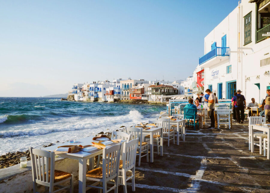 Charming waterfront dining with white tables, Greek architecture, and serene seaside views.