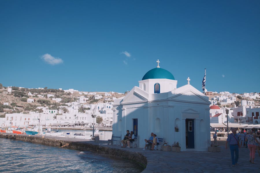 Charming Mediterranean coastal scene with white chapel, blue domes, boats, and hillside buildings.