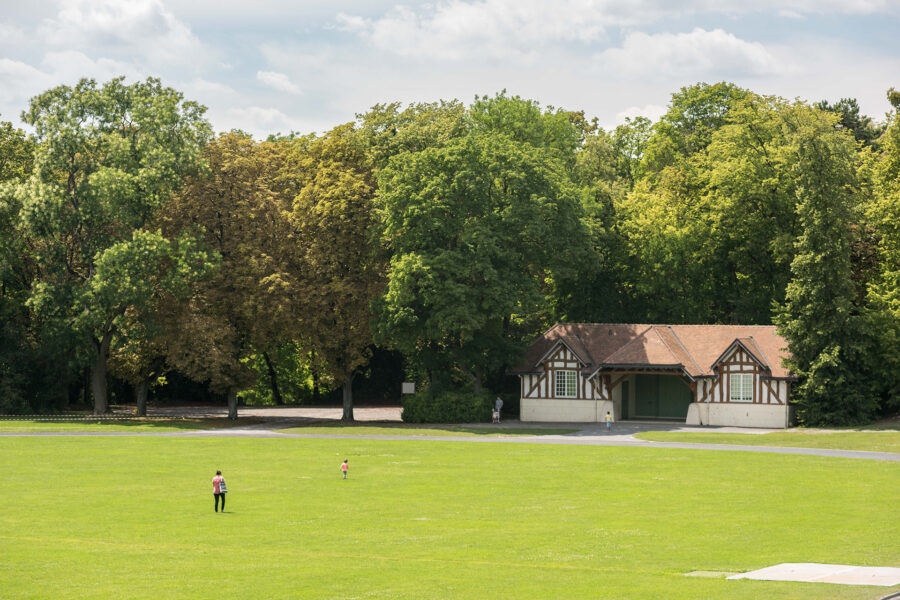 Green lawn park with timber pavilion, mature trees, and people strolling on a sunny day.