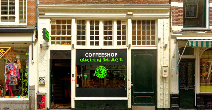 Coffeeshop storefront named "Green Place" with green sign and logo, flanked by a clothing store with colorful displays on its left. Brick facade, urban setting.