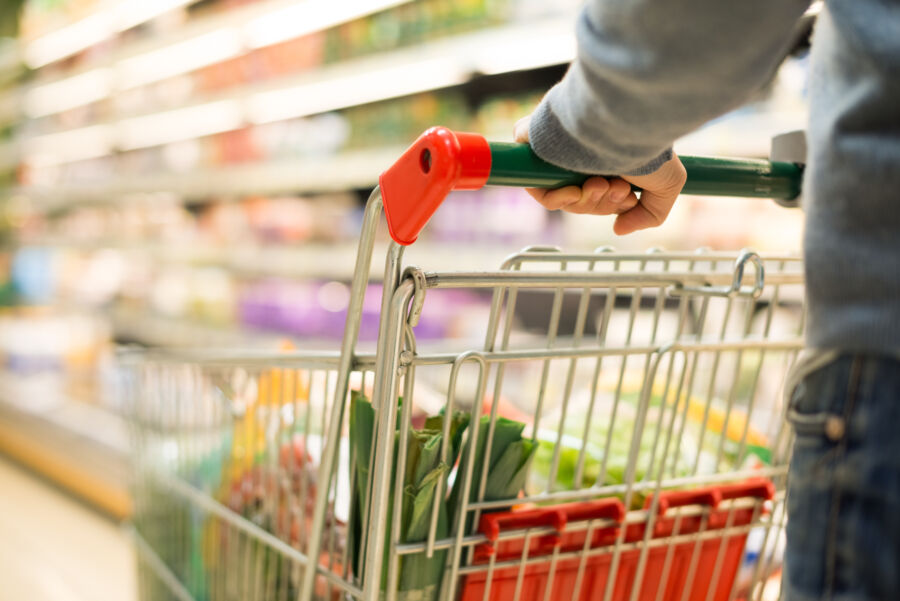 Person pushes a shopping cart filled with grocery items in a well-lit supermarket aisle. Bright shelves of products are blurred in the background.