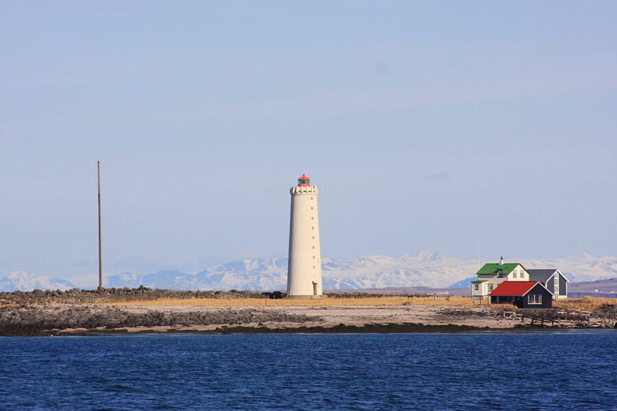 Grótta Lighthouse stands on the shore, overlooking a tranquil body of water under a clear blue sky.