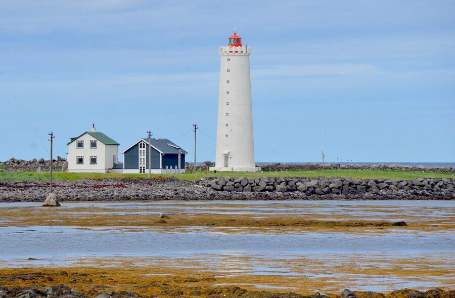 Grótta Lighthouse, a tall white structure with a red top, overlooks a rocky shore and two small houses under a blue sky.