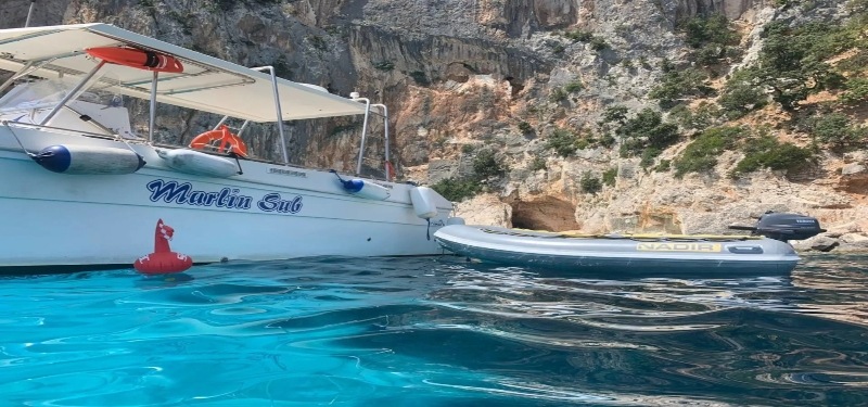 White boat with "Marlin Sub" on the side is anchored in clear turquoise water near rocky cliffs. A small, empty dinghy floats beside it.