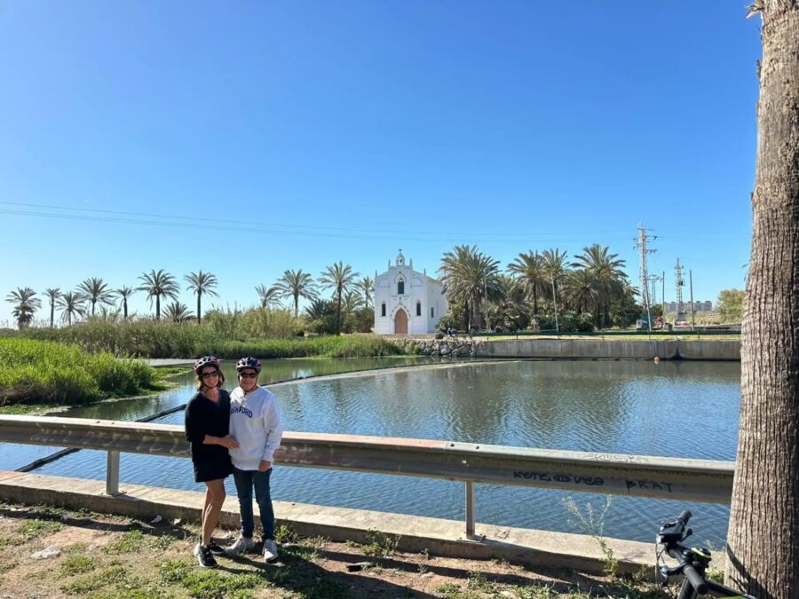 Couple in helmets stands by a tranquil pond, with a white church and palm trees under a clear blue sky in Valencia.