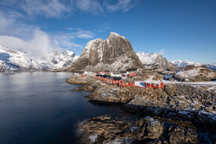 Sunny winter day in Hamnøy, Lofoten Islands, Norway, showcasing traditional red cabins against a snowy backdrop.
