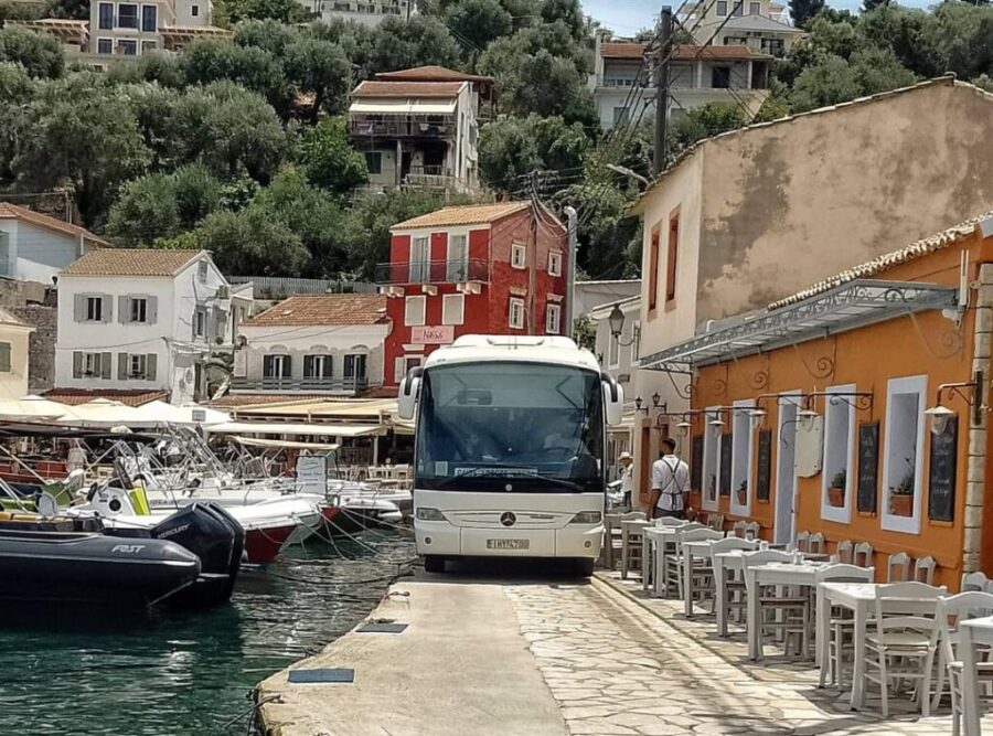 Coach bus parked on stone quay beside boats and waterfront cafe in coastal harbor town.