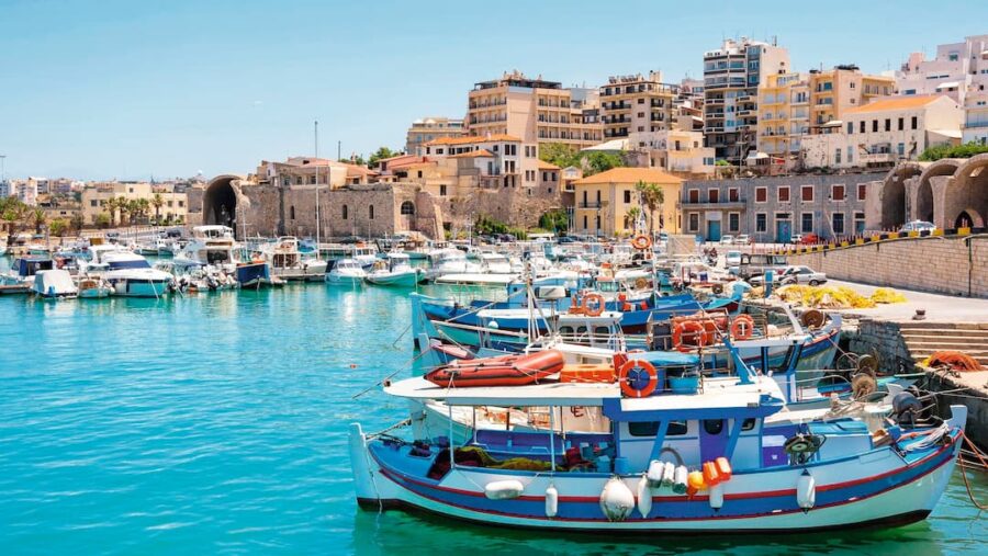 Sunlit boats in turquoise harbor, vibrant coastal architecture, lush greenery, clear skies.
