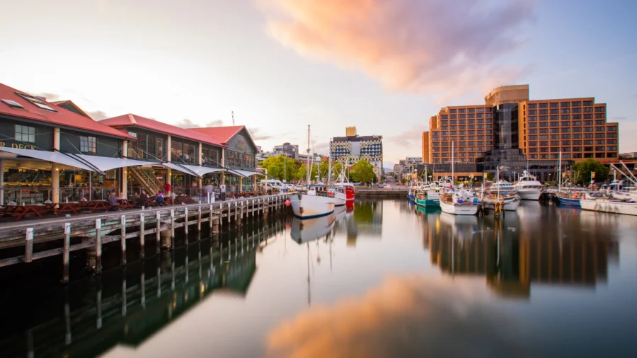 Waterfront harbor at sunset with boats, boardwalk restaurants and modern city buildings reflected in water.