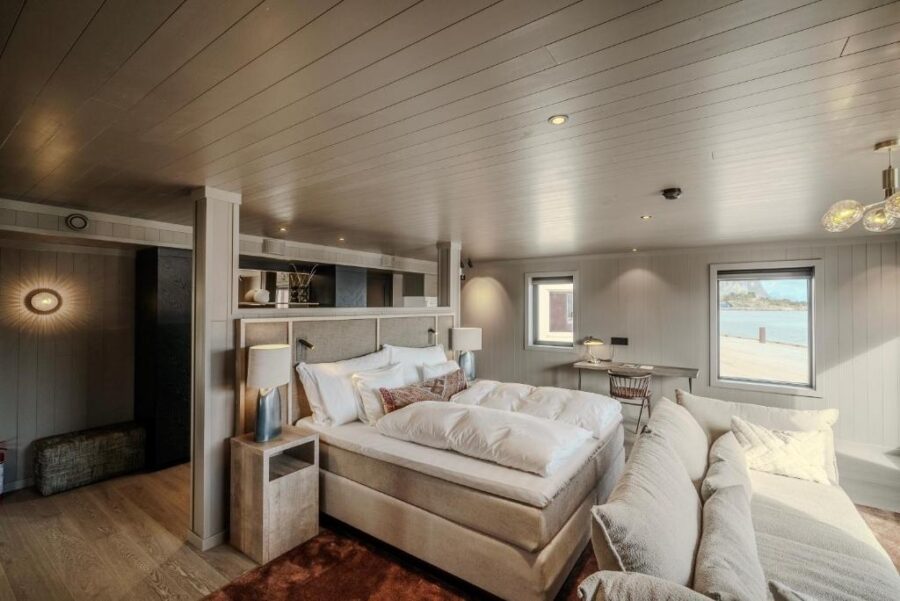 Cozy, modern bedroom with a large, plush bed, beige couch, and wooden ceiling of Henningsvær Bryggehotell - by Classic Norway Hotels.