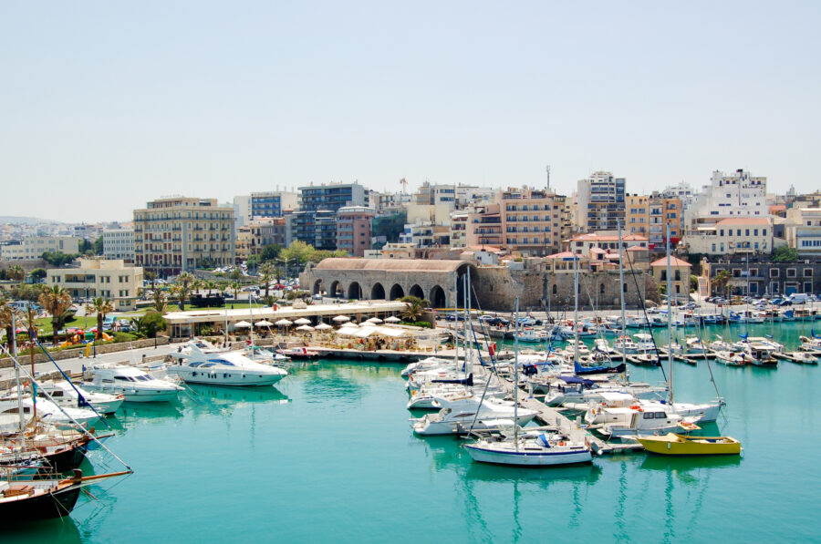 Scenic harbor in Heraklion, Crete, featuring boats, clear blue waters, and historic buildings along the waterfront.