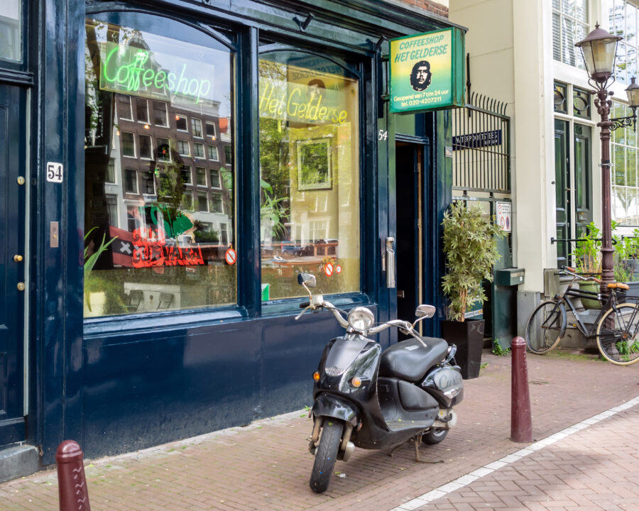 Front of a blue-painted coffeeshop with large windows, a scooter parked outside. Signs read "Coffeeshop Het Gelderse." A bicycle and plants are nearby.