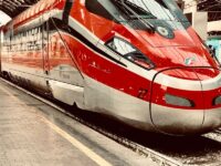 Red high-speed train at urban platform, sleek design, modern rail technology, passengers boarding.