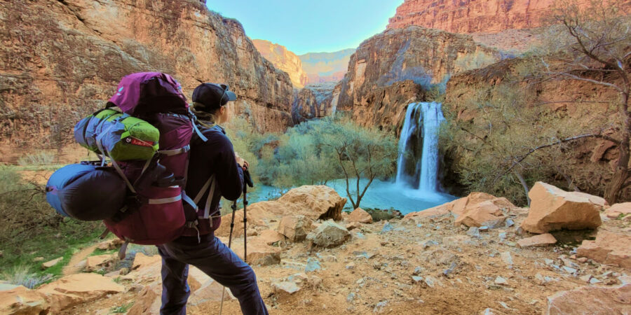 Hiker with backpack overlooks colorful canyon and waterfall, showcasing natures beauty.