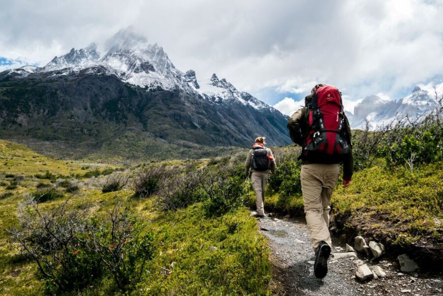 Hikers on narrow mountain trail, scenic snowy peaks and dynamic clouds in background.