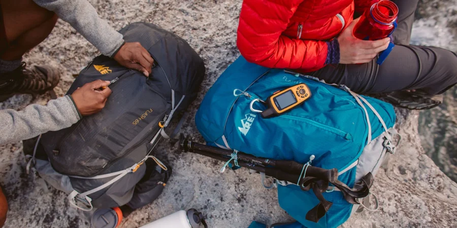Hikers with gear in rocky terrain: backpacks, tools, GPS, jackets, hydration, trekking poles.