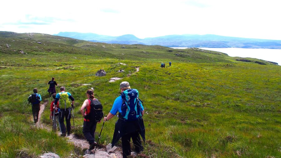 Hikers trekking through lush green hills with mountains and water views in Scotland.