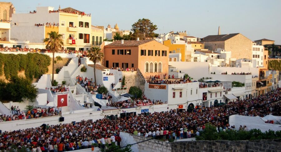 Sant Joan festival crowd on hillside terraces in sunlit Mediterranean town.