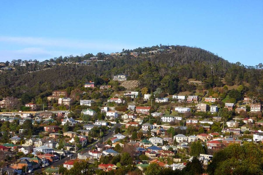 Hillside suburb in South Hobart with terraced homes, forested slopes, and a large hillside sign.