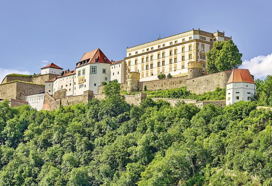 Historic Veste Oberhaus hilltop fortress with medieval stone walls and red roofs above lush green forest.