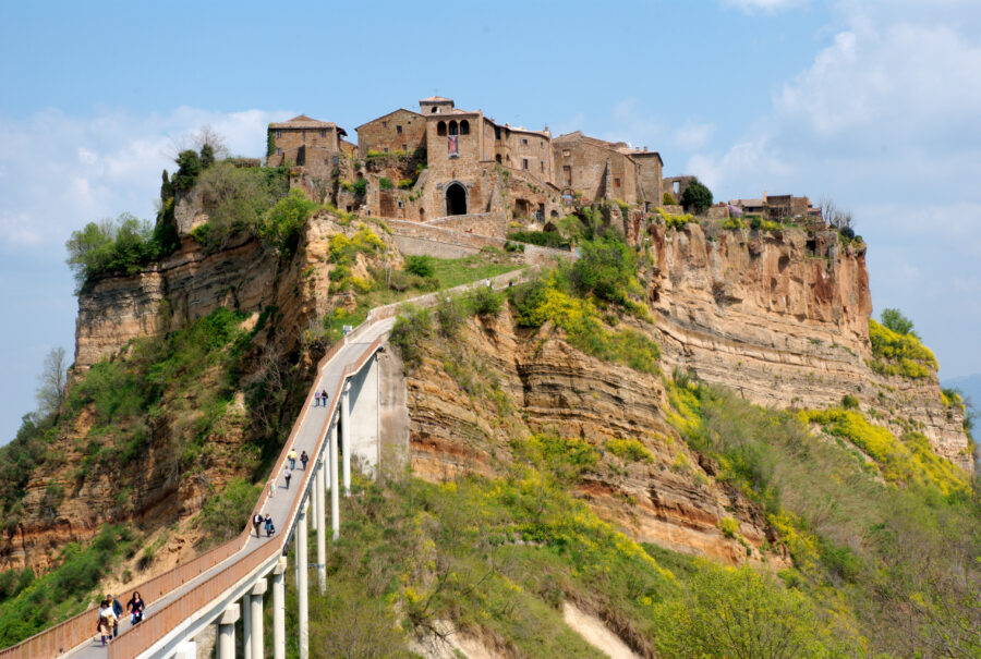 Medieval cliffside village with pedestrian bridge, stone buildings, and lush spring landscape.