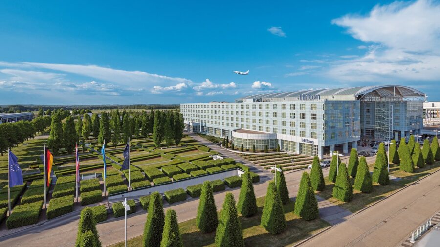 Modern Hilton Munich Airport hotel with glass facade, manicured gardens, and airplane overhead.