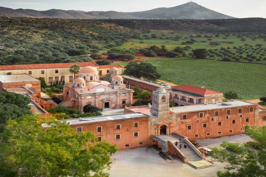 Historic complex amid lush landscape with rolling hills, mountain backdrop, vineyard, and olive trees.