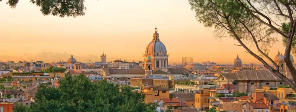Golden sunset over historic city with classical dome, terracotta rooftops, and trees.