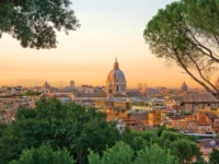 Golden sunset over historic city with classical dome, terracotta rooftops, and trees.