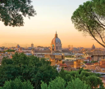 Golden sunset over historic city with classical dome, terracotta rooftops, and trees.