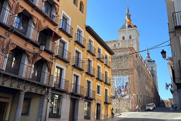 Toledo, Spain street with colorful buildings, stone architecture, and a tower adorned with a flag. The scene is bright and sunny.