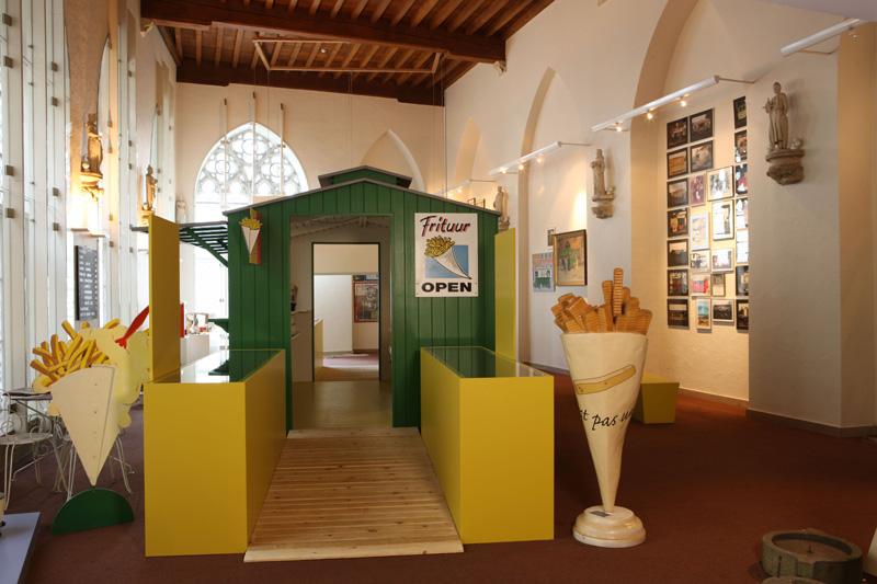 Historic hall museum installation with Frituur fries kiosk, oversized potato sculptures, and framed artworks.