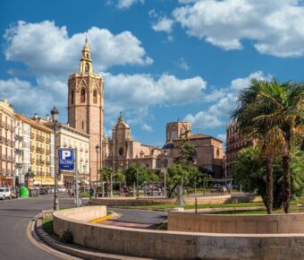 Vibrant Mediterranean city square with historical architecture, palm trees, and bustling street life.