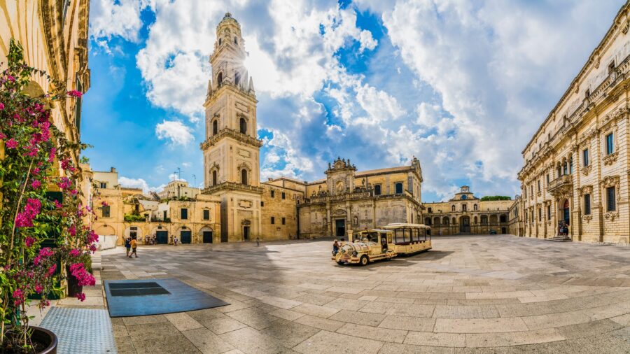 Vibrant historic square with tram, bell tower, floral display, and stone architecture in Lecce.