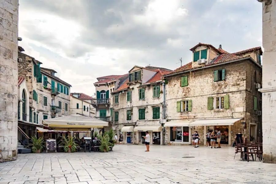 Charming historic square with stone buildings, green shutters, café, and social gathering.
