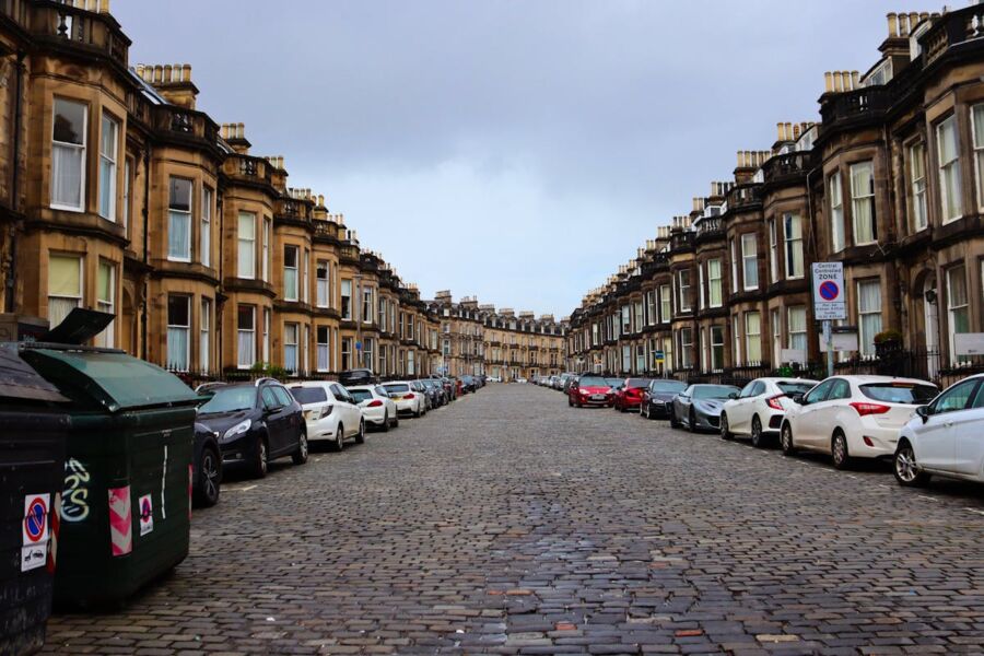 Victorian townhouses on cobblestone street with parked cars, cloudy skies, and urban elements.