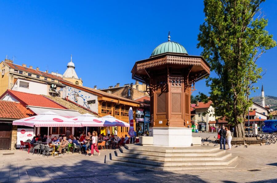 Historic Baščaršija square fountain with wooden pavilion, cafes, mosque minaret and surrounding hills.