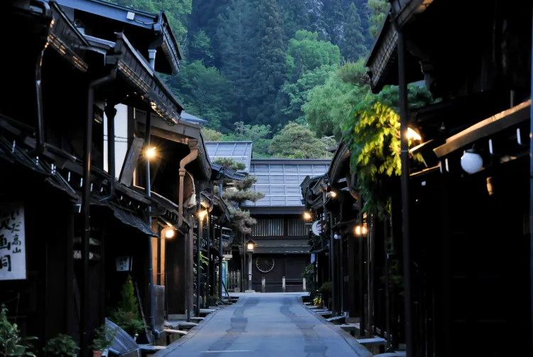Quiet traditional Japanese wooden street at dusk, empty road, lanterns glowing, mountains in background.