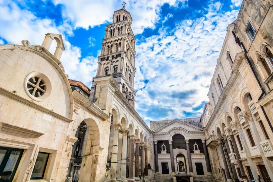 Historic European architecture with ornate bell tower and courtyard under a blue, clouded sky.