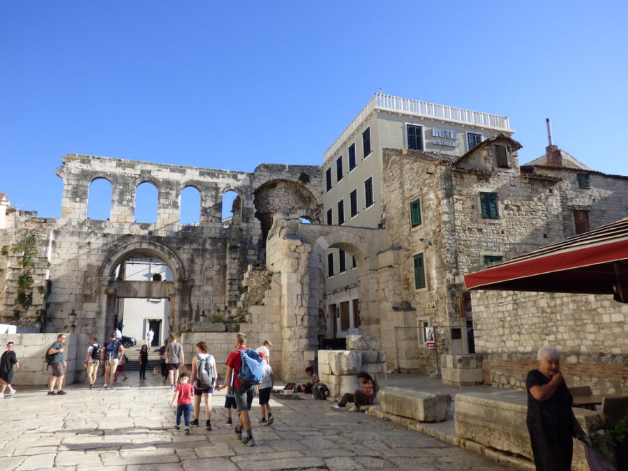 Ancient Diocletians Palace ruins with tourists, hotel, and café under vibrant blue sky.