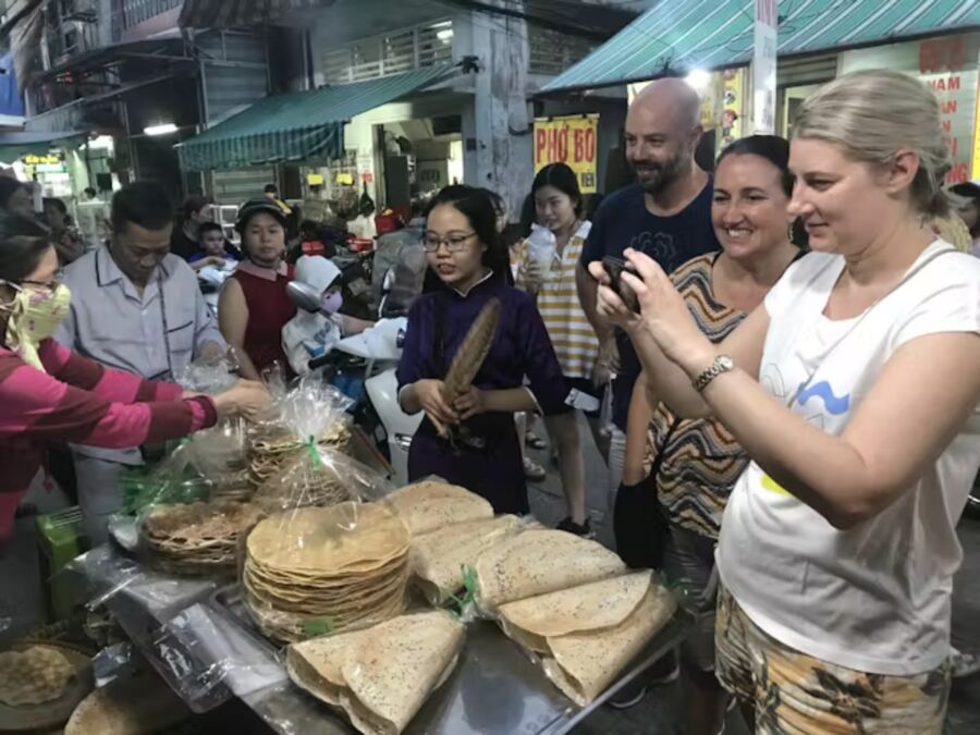 Participants of a Ho Chi Minh City food tour stand around a table, sharing and tasting local dishes at night.