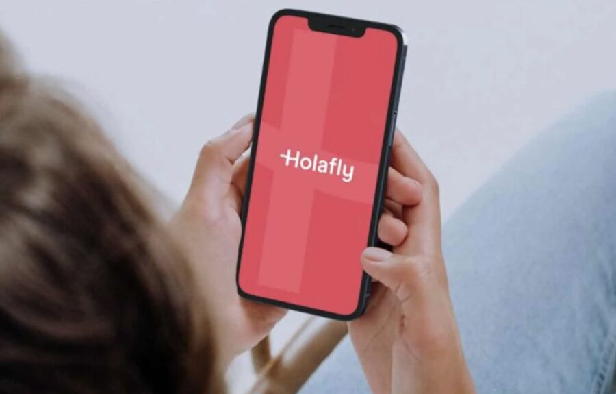 Person holding smartphone displaying Holafly app interface with red logo in cozy indoor setting.