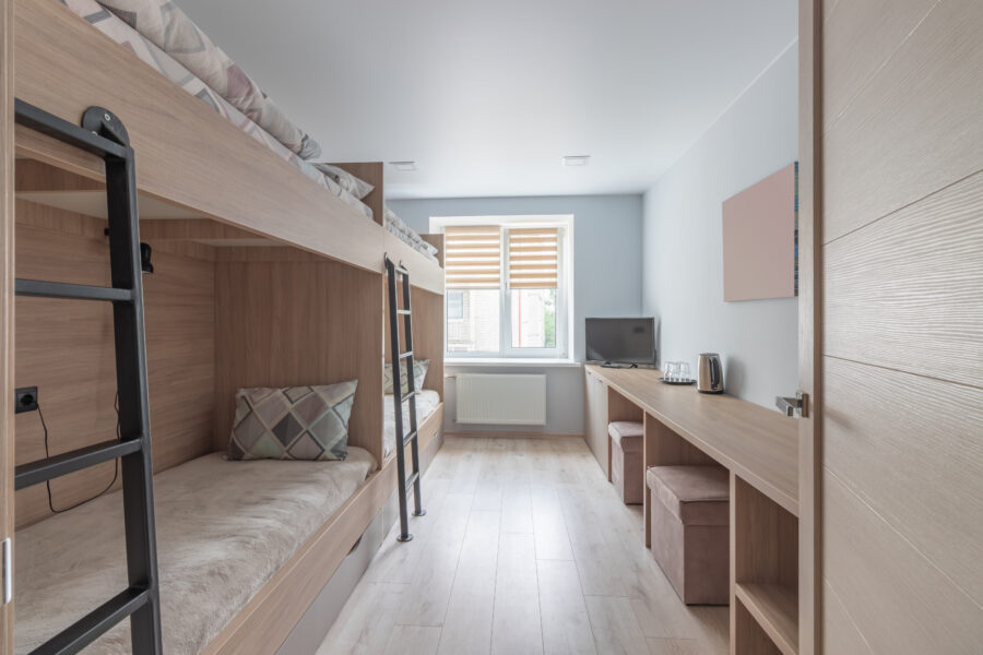 Modern dorm room with light wood bunk beds on the left and a long desk with a TV on the right. A window with blinds lets in natural light. Neutral tones.