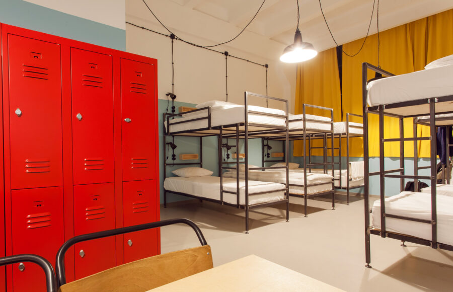 Modern hostel room with red lockers, metal bunk beds, and a warm atmosphere. Yellow curtains and an overhead lamp add a cozy touch.