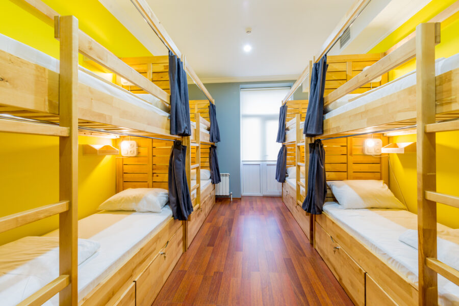 Bright hostel room with wooden bunk beds on each side, blue privacy curtains, and white bedding on hardwood floors against vibrant yellow walls. Cozy ambiance.