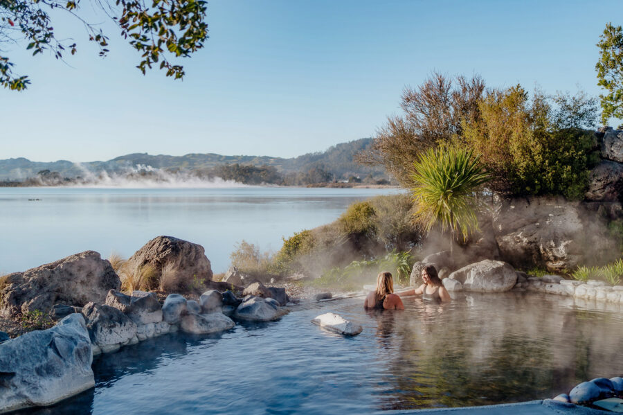 Lakeside geothermal hot spring pool with people relaxing in natural rock spa, steam, and mountain views.