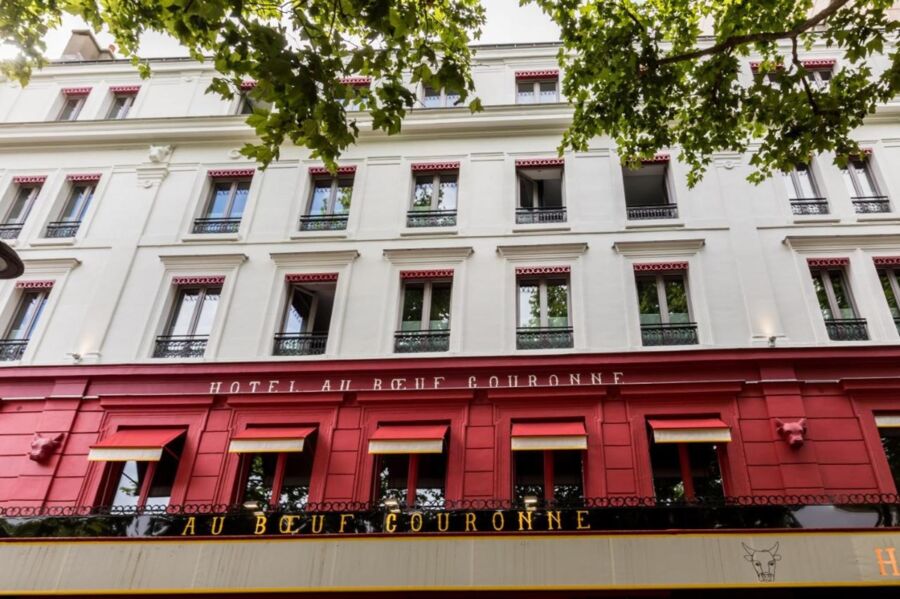 Facade of Hôtel Au Bœuf Couronné with red shutters and lush trees in urban setting.