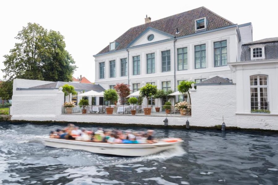 People enjoy a boat ride on a river by Hotel Van Cleef in Bruges, with picturesque surroundings.