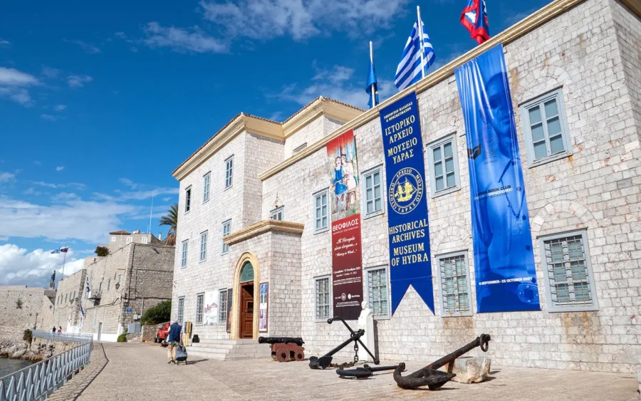 Hydra Historical Archives Museum on seafront with anchors, cannons, and waterfront promenade.
