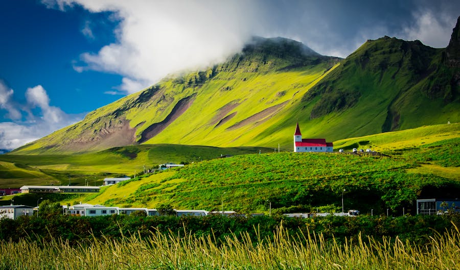 Icelandic landscape with green hills, mountains, and a red-spired white church under dramatic skies.